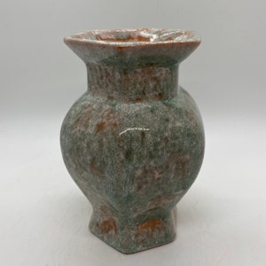 May include: A small, hexagonal ceramic vase with a green and brown glaze. The vase has a wide mouth and a rounded body.