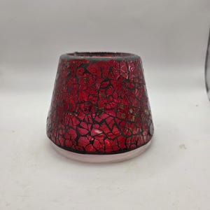 May include: A red mosaic glass candle holder with a cracked, textured appearance. The candle holder has a wide base and tapers upwards, with a dark rim. The base is a light pink colour.