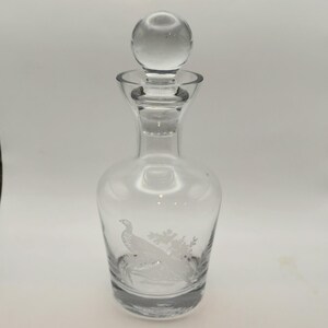 May include: Clear glass decanter with a round stopper. The decanter features an etched design of a bird and foliage. The stopper is a clear, round ball shape. The decanter is designed to hold liquids.