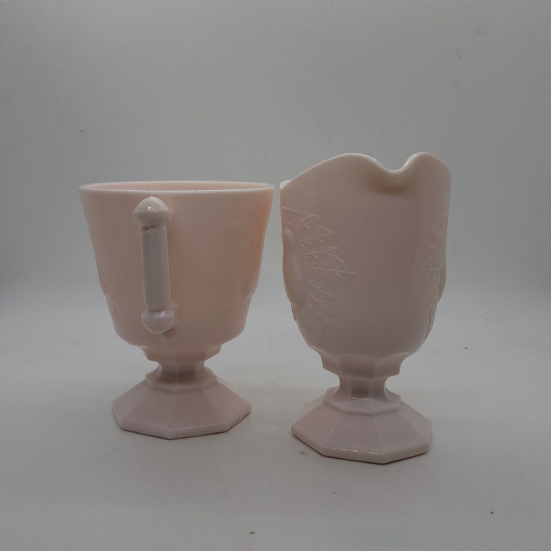 K&ouml;nnte beinhalten: Zwei passende rosa Glasserviceteile. Eines ist eine Tasse mit Henkel, das andere ein Sahnek&auml;nnchen mit gebogenem Ausguss. Beide Teile haben ein florales Bl&auml;ttermuster und stehen auf einem sechseckigen Sockel. Der Griff der Tasse ist wei&szlig;.