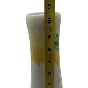 May include: A white ceramic vase with a yellow and green design. The vase is approximately 10 inches tall, as measured by a yellow measuring tape. The vase has a flared top and a slightly curved body.