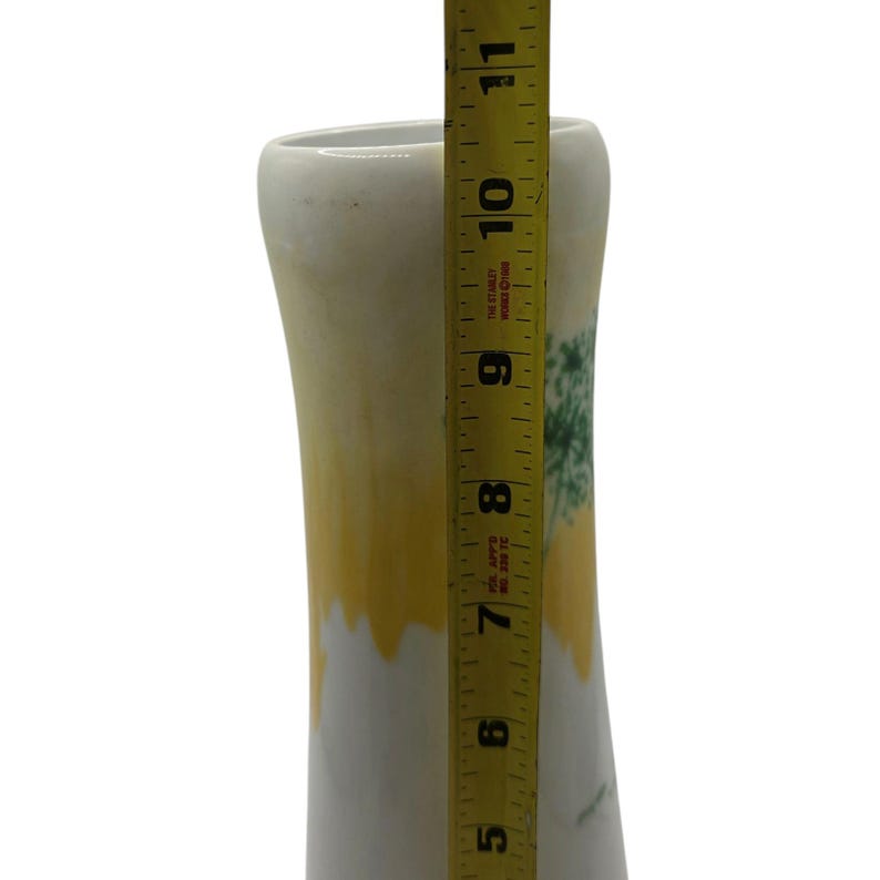 May include: A white ceramic vase with a yellow and green design. The vase is approximately 10 inches tall, as measured by a yellow measuring tape. The vase has a flared top and a slightly curved body.