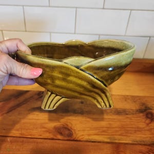 May include: A vintage, glazed ceramic bowl in shades of olive green and brown. The bowl has a unique, leaf-like design with three curved feet. The rim has a scalloped edge. The bowl is displayed on a wooden surface.