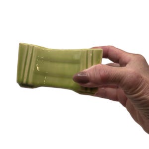 May include: A light green ceramic object with a rectangular, slightly concave shape and vertical ribbed detailing on both ends. The piece features a smooth, glossy finish and a solid, uniform color throughout its entire surface structure.