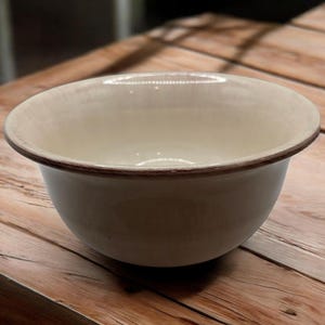 May include: A cream-colored ceramic bowl with a dark brown rim. The bowl has a rounded shape and sits on a wooden surface. The interior of the bowl is a lighter shade of cream.