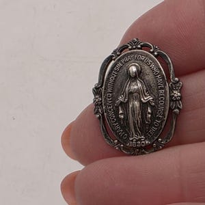 May include: A silver-toned oval religious pendant featuring an engraved image of a figure with outstretched arms. The pendant has ornate detailing around the edges and includes the text "O MARY CONCEIVED WITHOUT SIN PRAY FOR US WHO HAVE RECOURSE TO THEE".