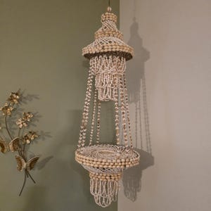 May include: A hanging tiered plant holder constructed entirely from small, off-white and tan seashells. The structure features a circular base platform suspended by multiple strands of shells, with decorative shell fringe at the top and bottom.
