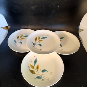May include: Four white ceramic dinner plates with a stylized leaf design in shades of teal, yellow, and brown. The plates have a black star pattern.