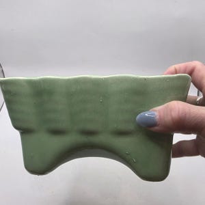 Könnte beinhalten: Hellgrüner Keramik-Pflanztopf mit rechteckiger Form und gewelltem oberen Rand. Der Pflanztopf hat ein einzigartiges Design mit drei gebogenen Beinen und einer glatten, glänzenden Oberfläche. Geeignet für kleine Pflanzen.