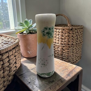 May include: A white ceramic vase with a yellow and green floral design. The vase has a cylindrical shape with a slightly flared top. The design features a dandelion motif. The vase is approximately 8 inches tall.