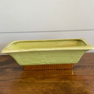 May include: A light green ceramic planter with a woodgrain pattern. The planter has a rectangular shape with a slightly flared lip and a brown base.