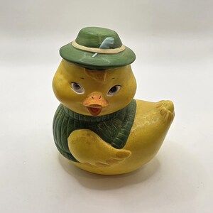 May include: A yellow ceramic duck figurine wearing a green hat and sweater. The duck has a detailed face with a bright orange beak and is posed in a seated position. The hat has a feather accent.