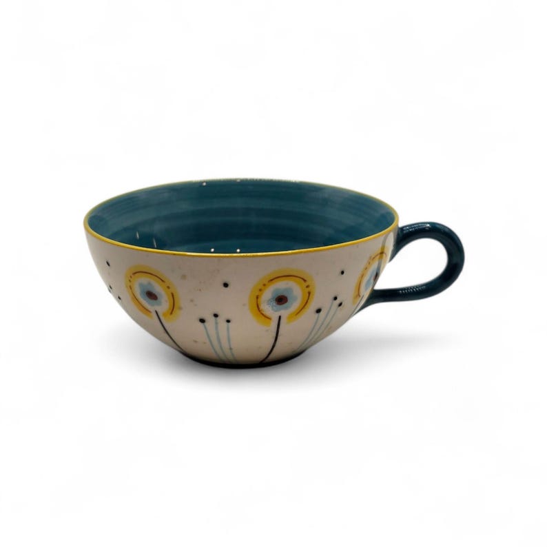 May include: Cream-colored ceramic soup bowl with a teal interior and handle. The exterior features a floral design with yellow and blue accents. The rim is outlined in yellow. The handle is teal.