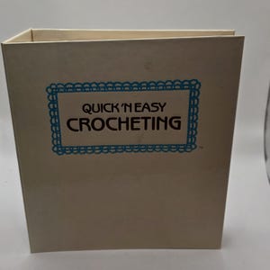 May include: A beige book with the title "Quick 'N Easy Crocheting" in black text, framed by a blue decorative border. The book is upright and appears to be a guide for crochet enthusiasts.