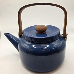 May include: A deep blue enameled metal teapot featuring a rounded body, a short spout, and a removable lid with a small, round wooden knob. The teapot includes a tall, arched wooden handle attached to the sides with metal hardware.