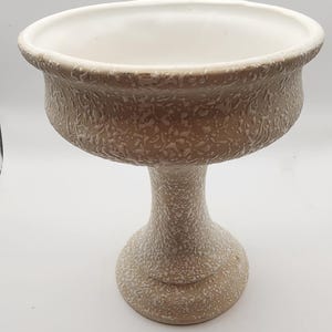 May include: A beige ceramic pedestal bowl with a textured surface. The bowl has a wide, shallow top with a white interior and a textured exterior. The pedestal base is a lighter beige color.