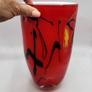 May include: A red glass vase with black and yellow abstract patterns. The vase has a wide body that narrows towards the base and a white rim. The black designs are painted on the surface, giving it a unique artistic appearance. The vase is approximately 25 cm tall.