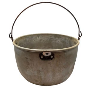 May include: A weathered, round metal pot with a dull silver-gray finish, featuring a thin, dark, arched wire handle attached to the sides and a small, protruding metal tab with a hole centered on the upper front rim of the vessel.