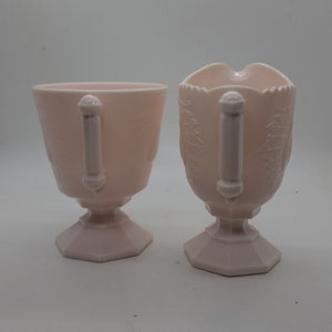 K&ouml;nnte beinhalten: Zwei passende Servierteile aus rosa Glas. Eines ist eine Tasse, das andere ein Sahnek&auml;nnchen mit Ausguss. Beide haben Griffe und sind mit einem Bl&auml;ttermuster verziert. Die Tasse und das Sahnek&auml;nnchen stehen auf einem Sockel.