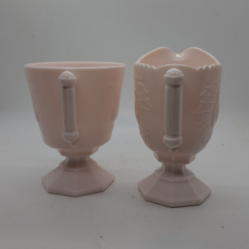K&ouml;nnte beinhalten: Zwei passende Servierteile aus rosa Glas. Eines ist eine Tasse, das andere ein Sahnek&auml;nnchen mit Ausguss. Beide haben Griffe und sind mit einem Bl&auml;ttermuster verziert. Die Tasse und das Sahnek&auml;nnchen stehen auf einem Sockel.