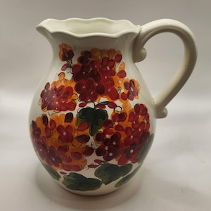 May include: Cream-colored ceramic pitcher with a large handle and a ruffled rim. The pitcher is decorated with a floral design in shades of red, orange, and green. The flowers are clustered around the body of the pitcher.