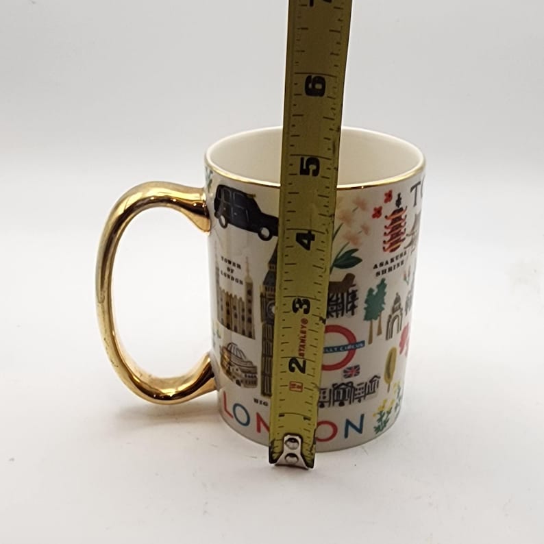 May include: A white ceramic mug with a gold handle and rim, featuring London landmarks and the word "LONDON". The mug is approximately 4 inches tall, perfect for coffee or tea.