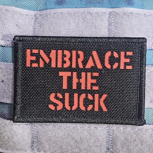 Pode incluir: Remendo retangular preto com as palavras "EMBRACE THE SUCK" em letras de bloco laranja. O remendo está sobre um fundo de tecido azul e cinza com tiras de velcro brancas.