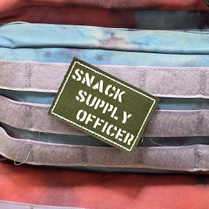 May include: A green patch with the text "SNACK SUPPLY OFFICER" in white lettering is attached to a backpack. The patch is rectangular with a white border. The backpack has a tie-dye pattern in shades of blue, pink, and purple.