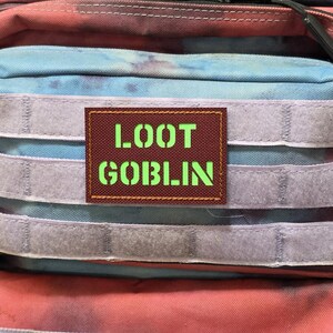 May include: A patch with the words "LOOT GOBLIN" in bright green letters on a brown background. The patch is rectangular and is attached to a backpack with blue, red, and purple fabric and gray velcro straps.