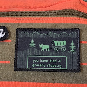 May include: A rectangular patch with a black border attached to an orange and olive green fabric surface. The patch features a dark green graphic of a covered wagon pulled by an ox, pine trees, mountains, and the text you have died of grocery shopping.