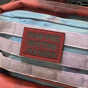 May include: A close-up of a multi-coloured bag with a burgundy patch. The patch has the words "SEMI-PRO BUTTON MASHER" in dark green. The bag has light blue, red, and purple accents.
