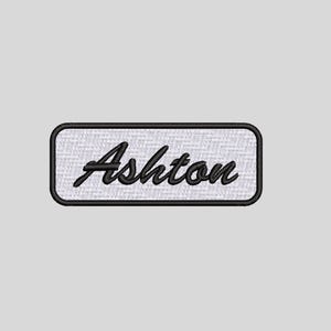 May include: A rectangular embroidered patch featuring a white background with a black border. The name Ashton is stitched in the centre in a black cursive script font. The patch has a clean, finished edge and a textured fabric appearance.