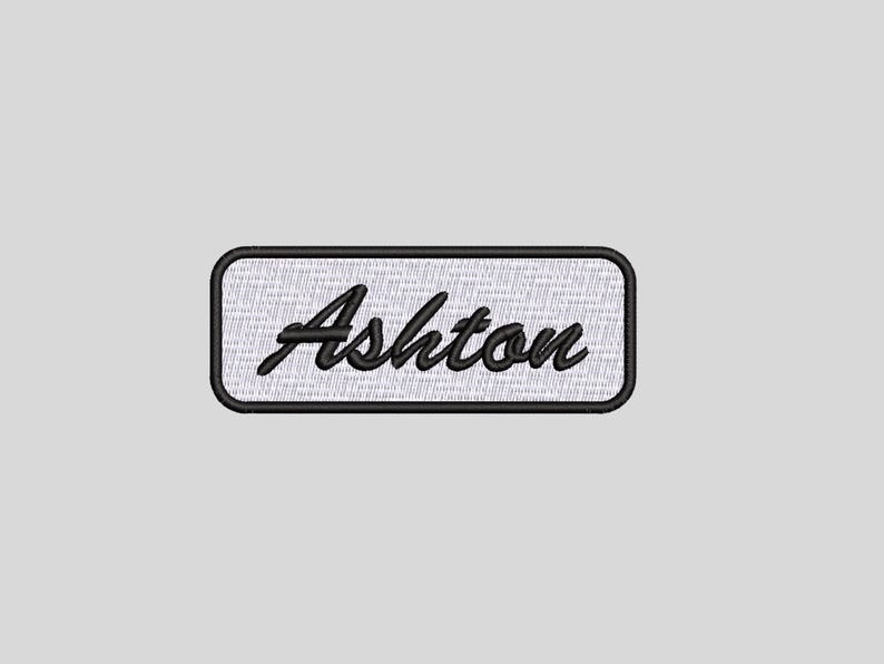 May include: A rectangular embroidered patch featuring a white background with a black border. The name Ashton is stitched in the centre in a black cursive script font. The patch has a clean, finished edge and a textured fabric appearance.