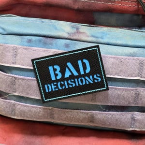 May include: A rectangular black patch with the words "BAD DECISIONS" in bright blue. The patch has a light blue border and is set against a backdrop of a tie-dye pattern in shades of blue, red, and purple.