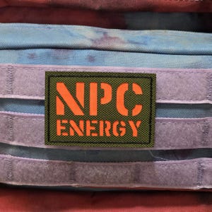 May include: A rectangular patch with the words "NPC ENERGY" in orange on a green background. The patch has a black border. The background features a tie-dye pattern in shades of red, blue, and purple.