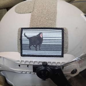 May include: A rectangular morale patch with a black border attached to a tan tactical helmet. The patch features a black cat with an open mouth against a background of repeating lowercase letter a characters in black and white horizontal stripes.