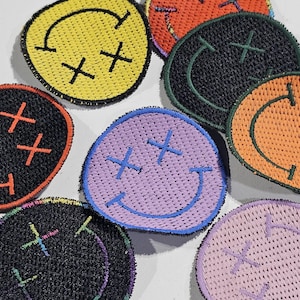 Puede incluir: Una colección de nueve parches bordados con caritas sonrientes en varios colores, incluyendo negro, amarillo, naranja, morado y verde. Cada parche presenta un diseño clásico de carita sonriente con dos ojos tachados.