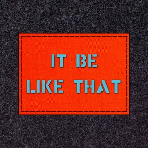 May include: An orange rectangular patch with the text "IT BE LIKE THAT" in light blue. The patch has a black stitched border and is set against a dark grey, textured background.