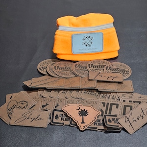 May include: A collection of brown leather patches with various designs and text, including "Old Plum", "ELS Kennels", "Skyla", "Tripp", "Frost", and "Reel Cool" along with a bright orange fleece neck warmer with a gray reflective strip and a logo that says "fourkeyslic.com".