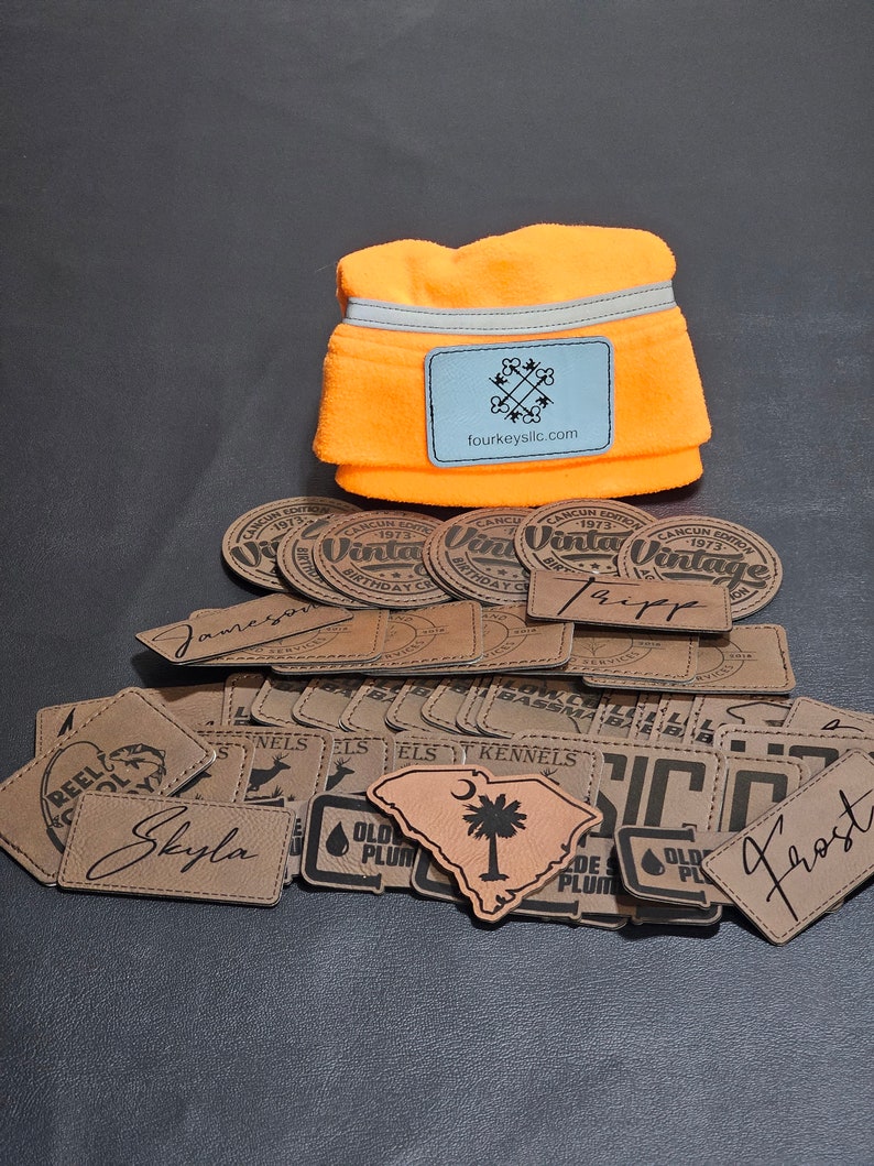 May include: A collection of brown leather patches with various designs and text, including "Old Plum", "ELS Kennels", "Skyla", "Tripp", "Frost", and "Reel Cool" along with a bright orange fleece neck warmer with a gray reflective strip and a logo that says "fourkeyslic.com".