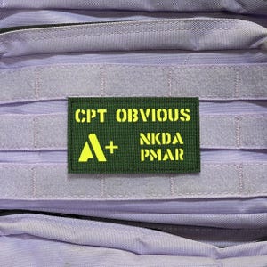 May include: A green patch with yellow text reading "CPT OBVIOUS", "A+", "NKDA PMAR". The patch is attached to a lilac-colored tactical bag, with visible webbing and Velcro straps. The text is in a bold, sans-serif font.