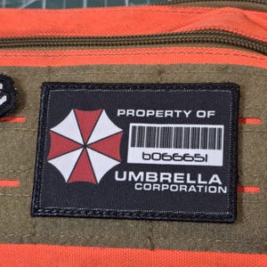 Op de afbeelding: Een rechthoekige zwarte patch met een rood-witte paraplu-afbeelding, een streepjescode en de tekst PROPERTY OF UMBRELLA CORPORATION b066651. De patch is bevestigd op een oranje en olijfgroene stoffen ondergrond met een zichtbare rits.