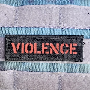 May include: A black rectangular patch with the word "VIOLENCE" in orange, stencil-style lettering. The patch is sewn onto a background of light blue and purple fabric with gray velcro straps.