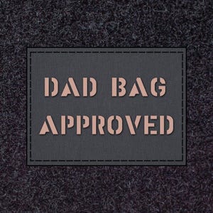 Puede incluir: Un parche gris oscuro con las palabras "DAD BAG APPROVED" en una fuente de plantilla marrón claro. El parche es rectangular con un borde cosido, sobre un fondo oscuro y texturizado.