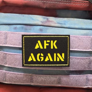 May include: A black patch with yellow text reading "AFK AGAIN." The rectangular patch has a yellow border and is attached to a multi-coloured fabric background. The background features shades of blue, purple, and red.