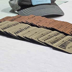 May include: A stack of brown leather patches with the text "The County Seat" printed in black. The patches are stacked on a white surface. A gray baseball cap with a blue patch that says "fourkeysllc.com" is in the background.