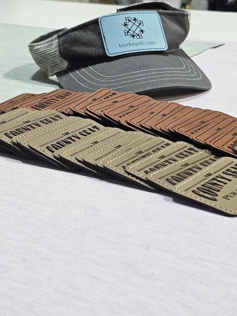 May include: A stack of brown leather patches with the text "The County Seat" printed in black. The patches are stacked on a white surface. A gray baseball cap with a blue patch that says "fourkeysllc.com" is in the background.