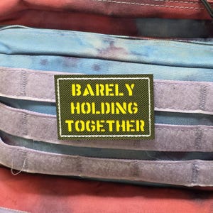 May include: A multi-coloured backpack featuring a patch with the text "BARELY HOLDING TOGETHER" in yellow on a green background. The backpack has blue, red, and purple sections with grey velcro straps.