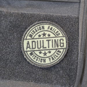 Op de afbeelding: Een ronde olijfgroene patch met een zwarte rand en zwarte geborduurde tekst die MISSION FAILED ADULTING MISSION FAILED leest. De patch bevat drie kleine sterren boven en onder het woord ADULTING en is bevestigd op een zwarte stof.
