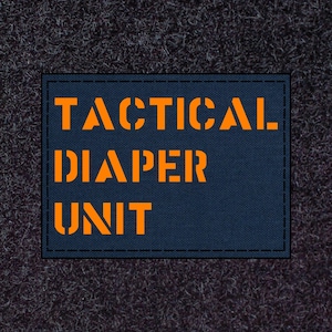 May include: A navy blue patch with the words "TACTICAL DIAPER UNIT" in orange, stencil-style lettering. The patch has a black border and is set against a dark gray, textured background.